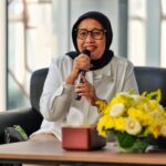 Woman wearing a black hijab and glasses speaks into a microphone during a panel with yellow flowers on the table nearby